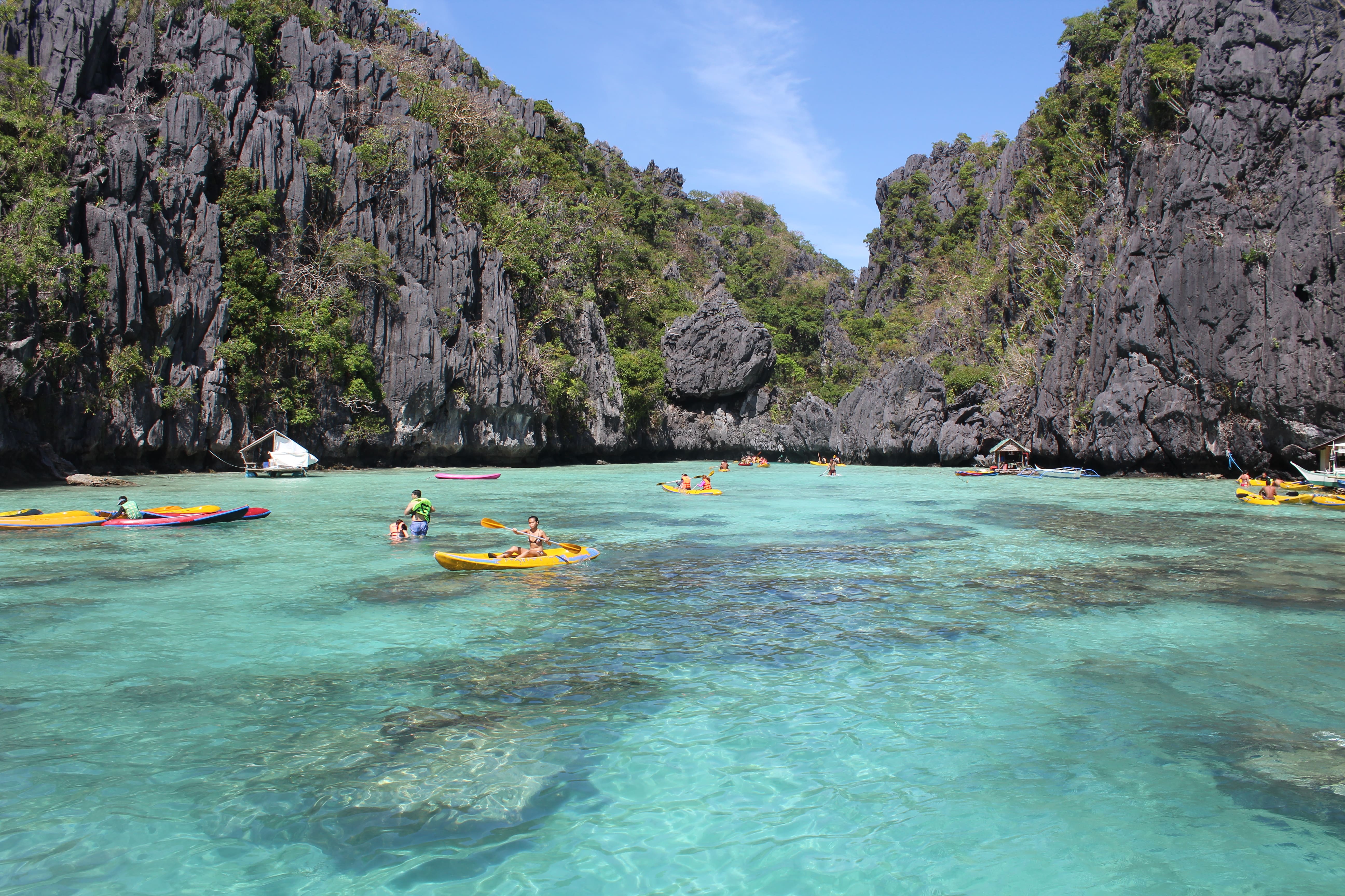 A vibrant tropical seascape featuring a clear turquoise lagoon surrounded by steep limestone cliffs covered in lush green vegetation. The water reveals underwater features like coral reefs or sandy areas. In the foreground, a yellow kayak with two people paddles through the water, while other colorful kayaks and swimmers enjoy the shallow areas. Small boats are anchored in the background near the cliffs. The sky is mostly clear with a few wispy clouds, and the overall scene is bright and lively, suggesting a popular spot for water activities.