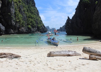 El Nido, Palawan with a white sandy beach, turquoise waters, limestone cliffs, an outrigger boat, and people swimming.