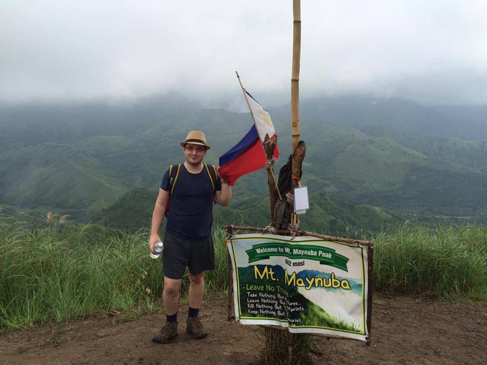A person with a backpack stands at Mount Maynoba summit, holding a Philippine flag, with green hills and a cloudy sky in the background, and a 'Welcome to Mt. Maynuba' sign.