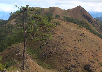 Mount Ulap with hikers on a trail, grassy hills, dense vegetation, and layered mountains under a cloudy sky with sunlight.