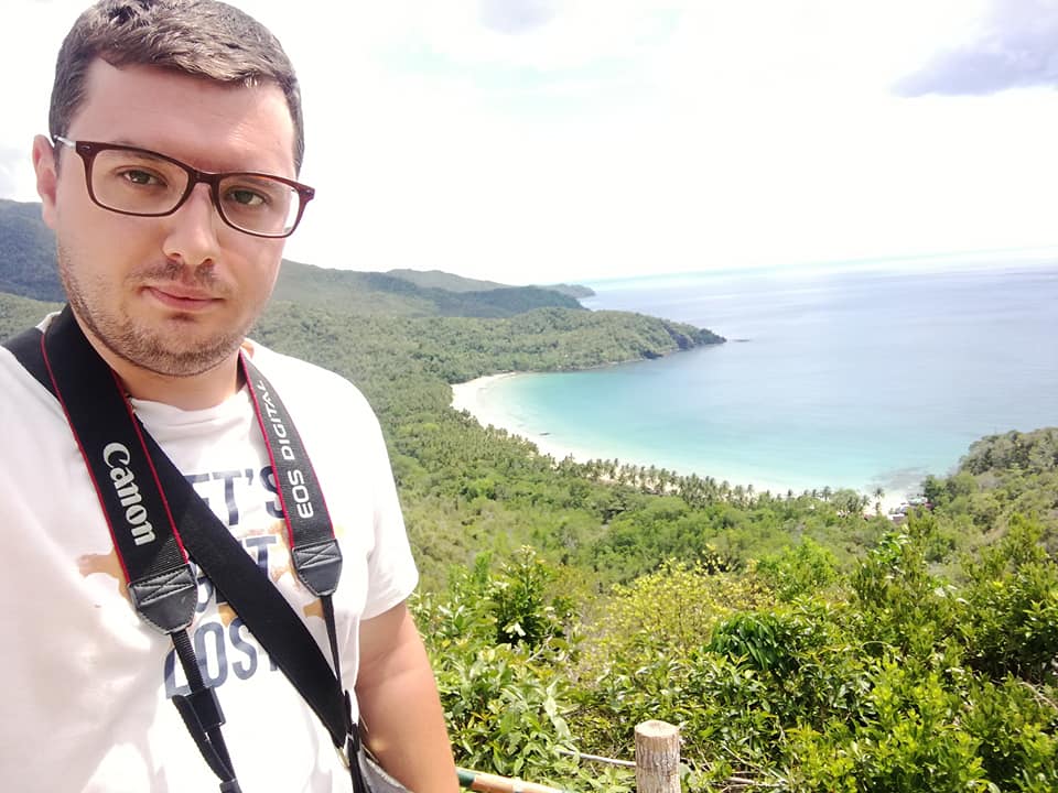 A person with a Canon camera strap stands in front of Nagtabon Beach, featuring turquoise waters, sandy shores, and green hills under a cloudy sky.