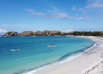 Tanjung Aan Beach in Lombok with white sand, turquoise waters, small boats, and rugged hills under a blue sky with clouds.