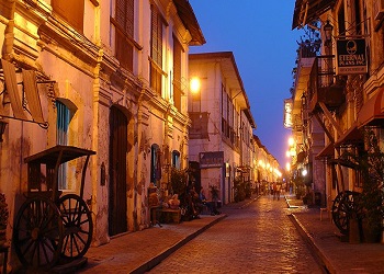Vigan City street with historic colonial buildings, cobblestone path, capiz windows, wooden cart, and warm street lights at dusk.