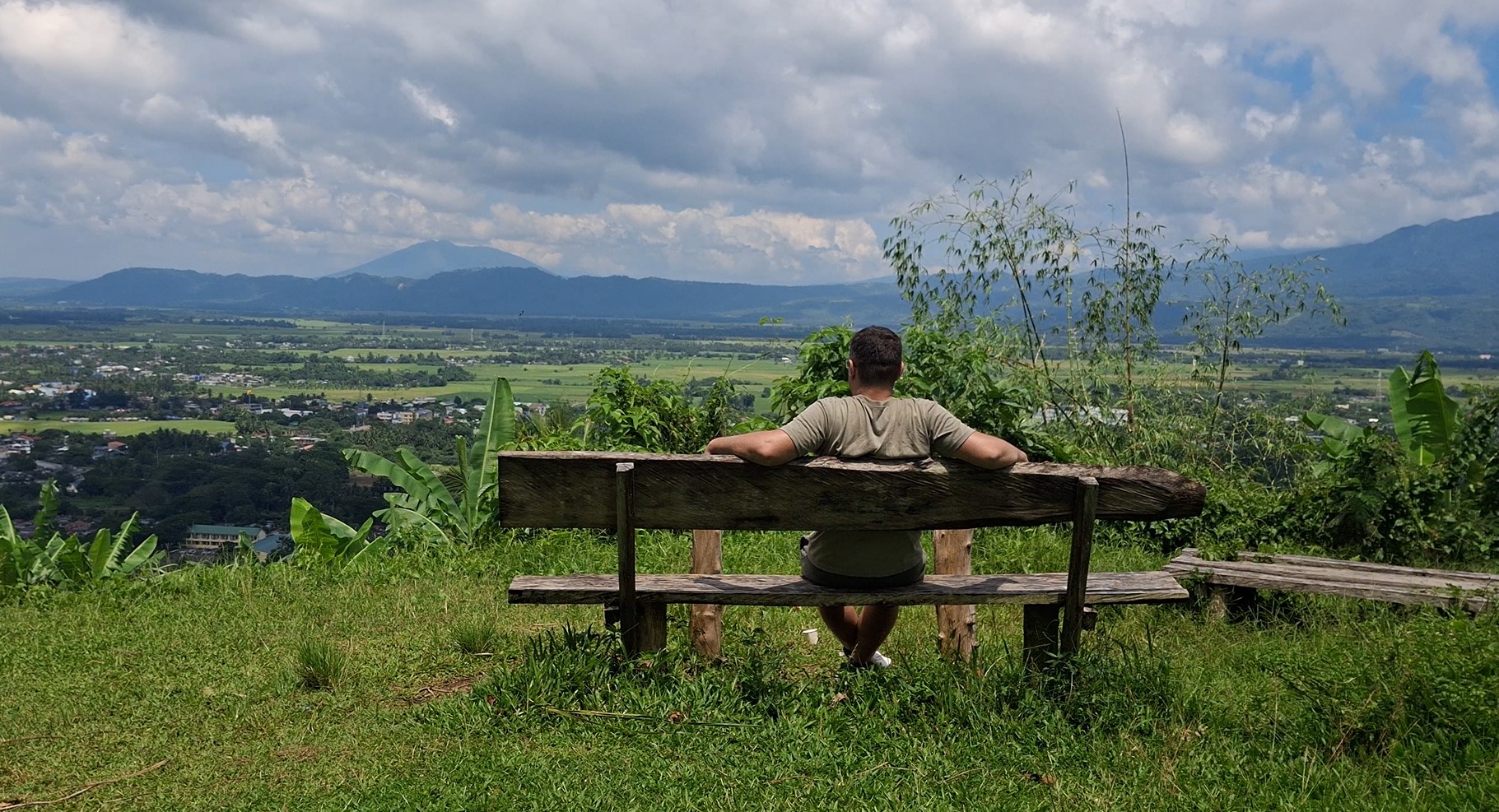 The Lazy Traveler sitting on a rustic wooden bench overlooking the vast green plains of Ligao City