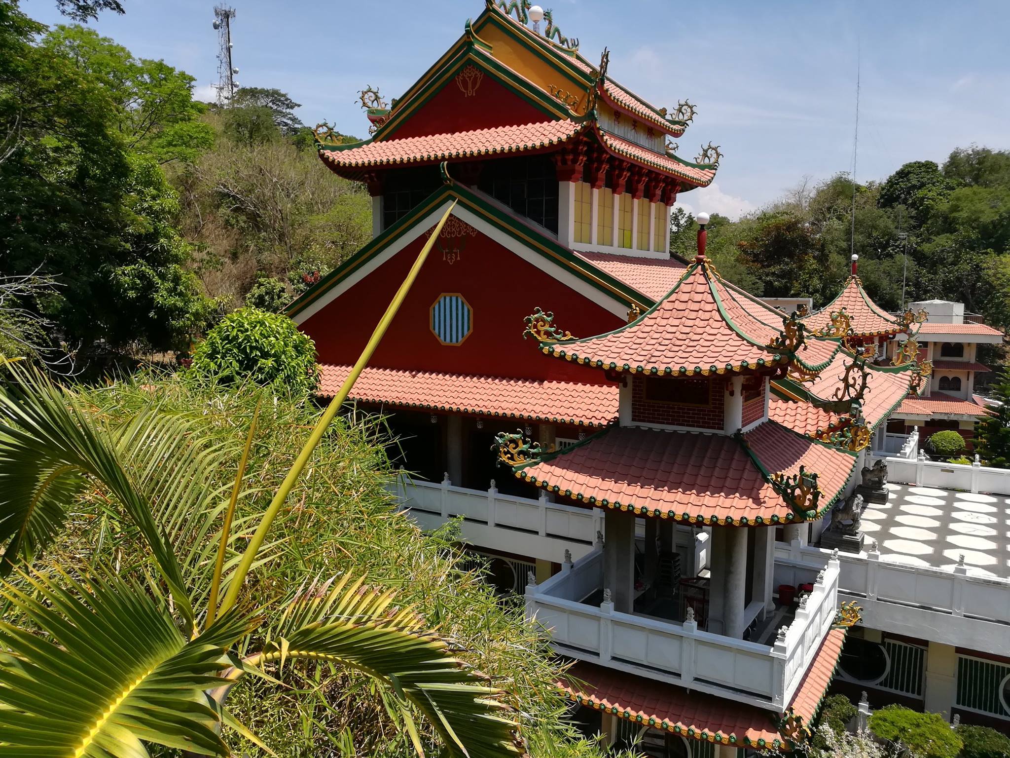 The colorful pagoda-style structure of Ma-Cho Temple, featuring red walls, green tiled roofs, and traditional Chinese decorations, nestled among dense trees and overlooking a hillside.