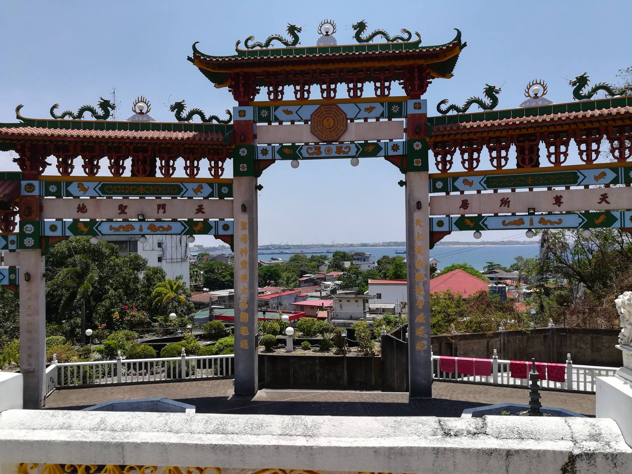 A vibrant, ornate gate of the Ma-Cho Temple in La Union, Philippines, featuring colorful dragons, intricate designs, and Chinese characters, with a scenic view of the ocean and town in the background.