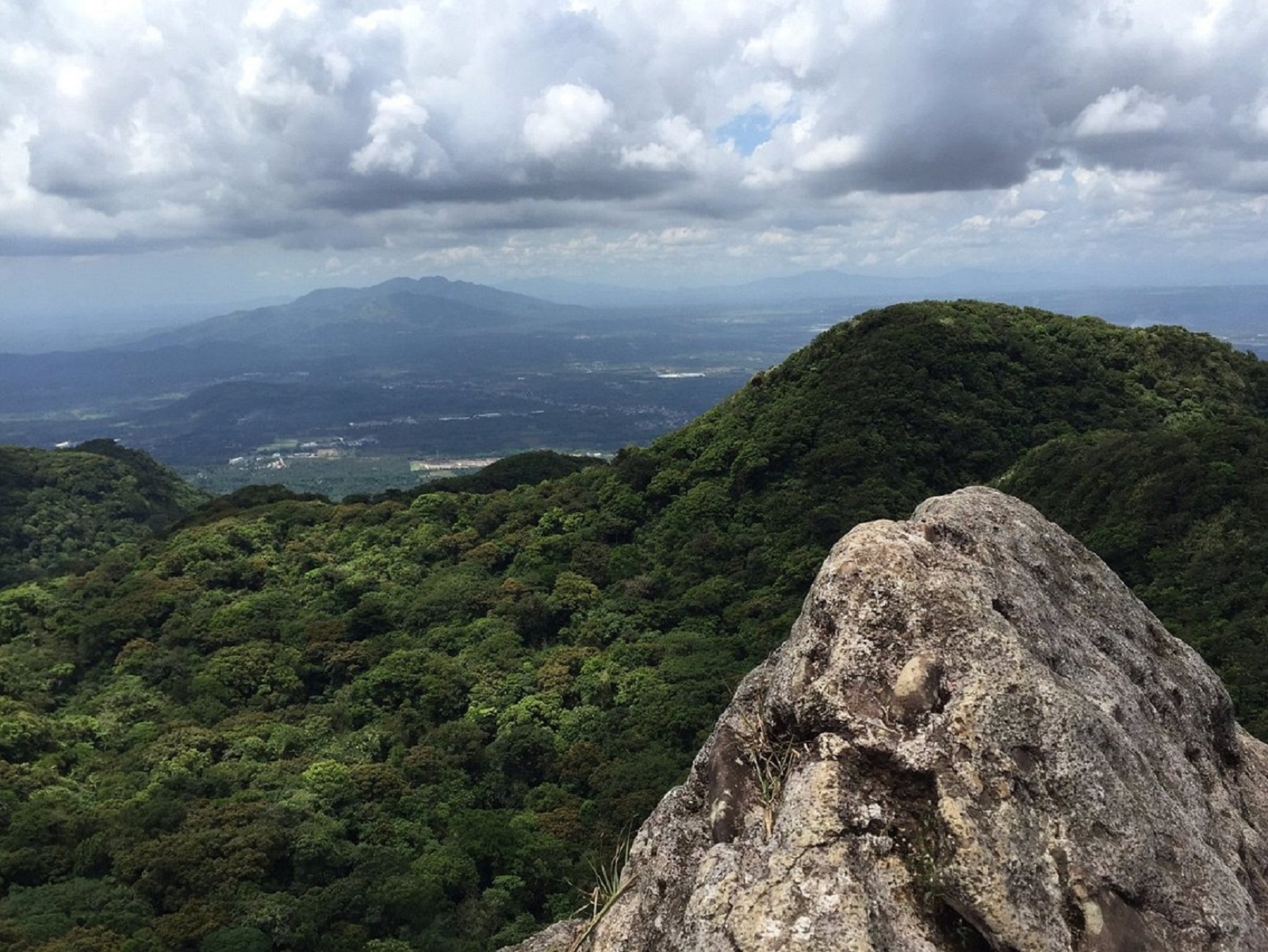 Panoramic view from a rocky outcrop on Mount Makiling, showcasing lush green forested hills, distant mountains, and a cloudy sky over the Philippines’ landscape.