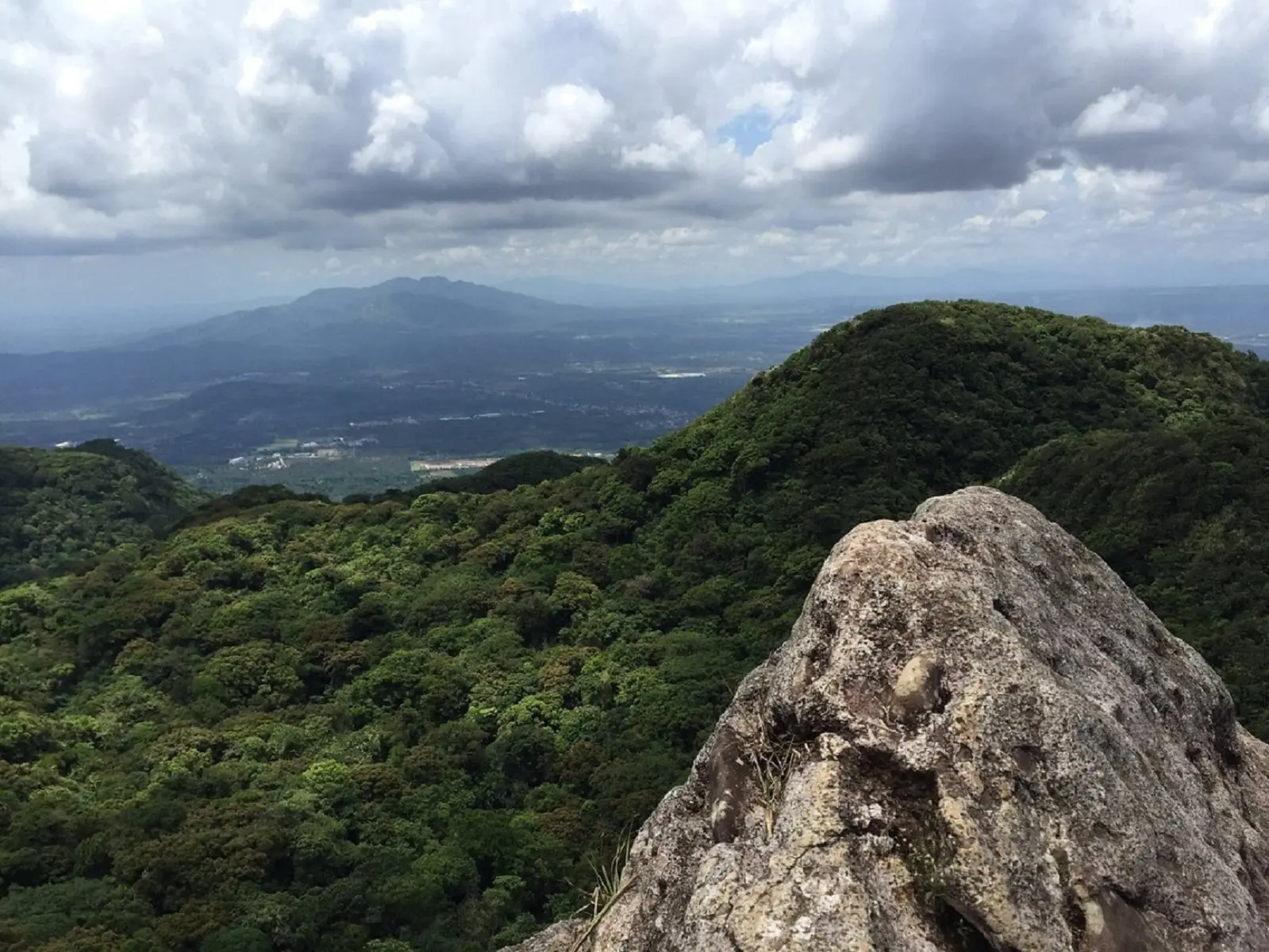 Panoramic view from a rocky outcrop on Mount Makiling, showcasing lush green forested hills, distant mountains, and a cloudy sky over the Philippines' landscape.