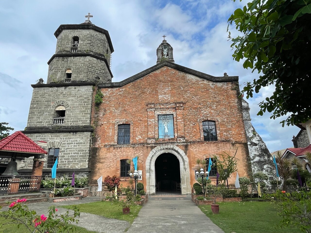Historic Boac Cathedral in Marinduque, Philippines, featuring a rustic brick facade, a tall stone bell tower, and an arched entrance, surrounded by lush greenery and colorful flags under a partly cloudy sky.