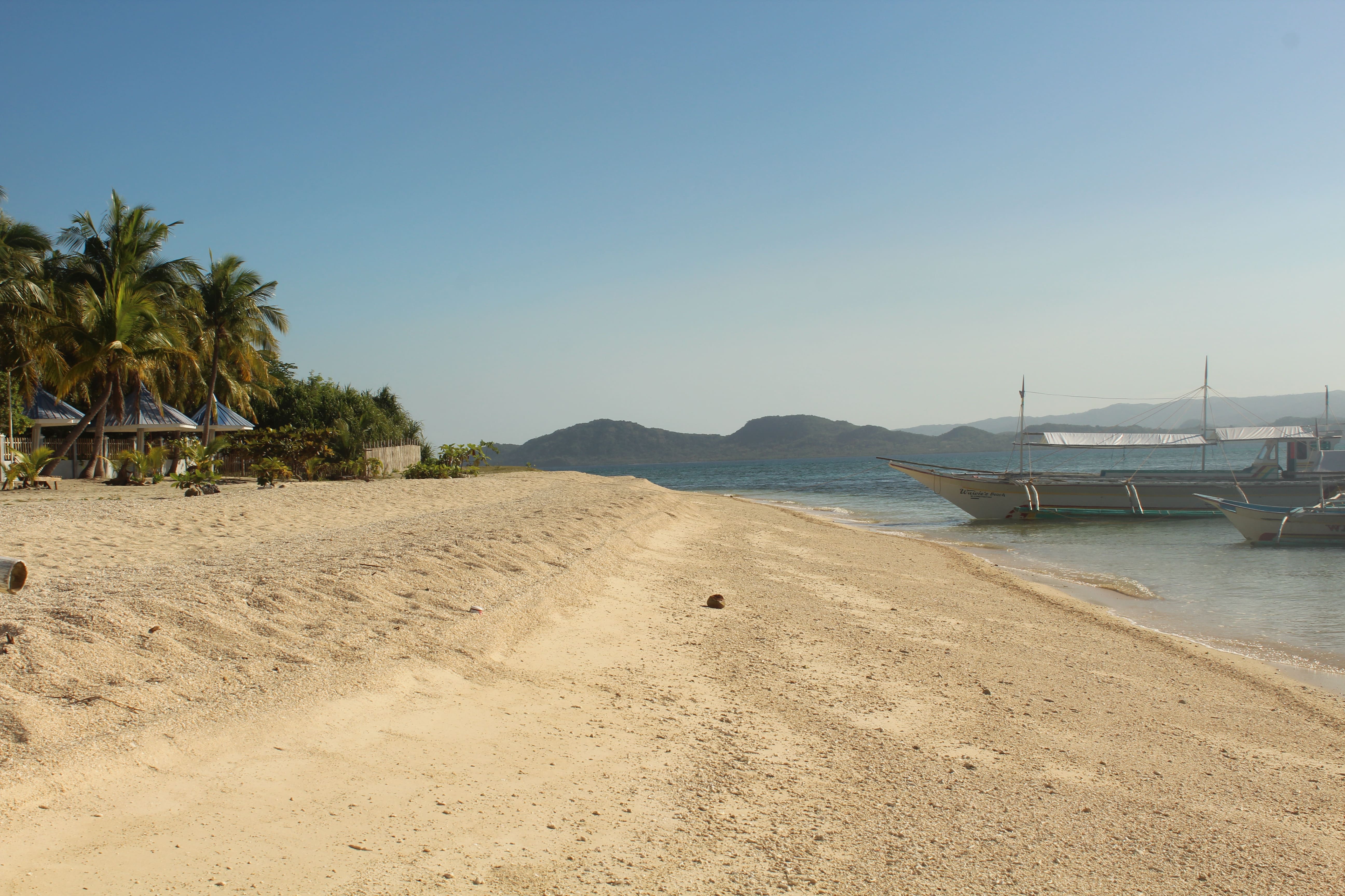 Scenic view of Maniwaya Island, a short boat ride from Marinduque, Philippines, featuring a pristine white sandy beach, calm turquoise waters, palm trees, traditional huts, and outrigger boats anchored near the shore, with distant green hills under a clear blue sky.