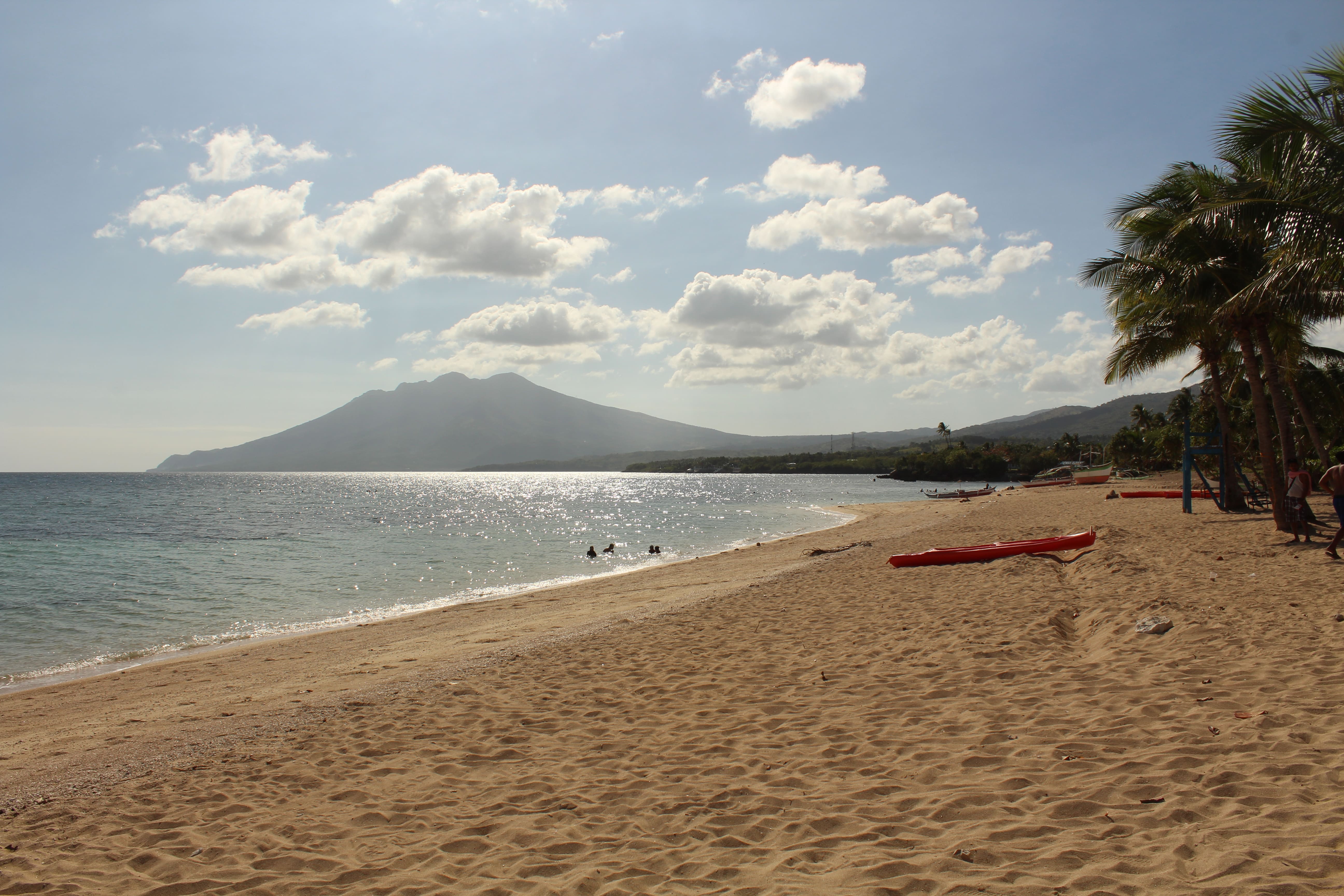 Scenic view of Poctoy Beach in Marinduque, Philippines, featuring golden sands, clear blue waters, palm trees, and a distant mountain under a bright sky with scattered clouds, with people enjoying the beach and red kayaks on the shore.