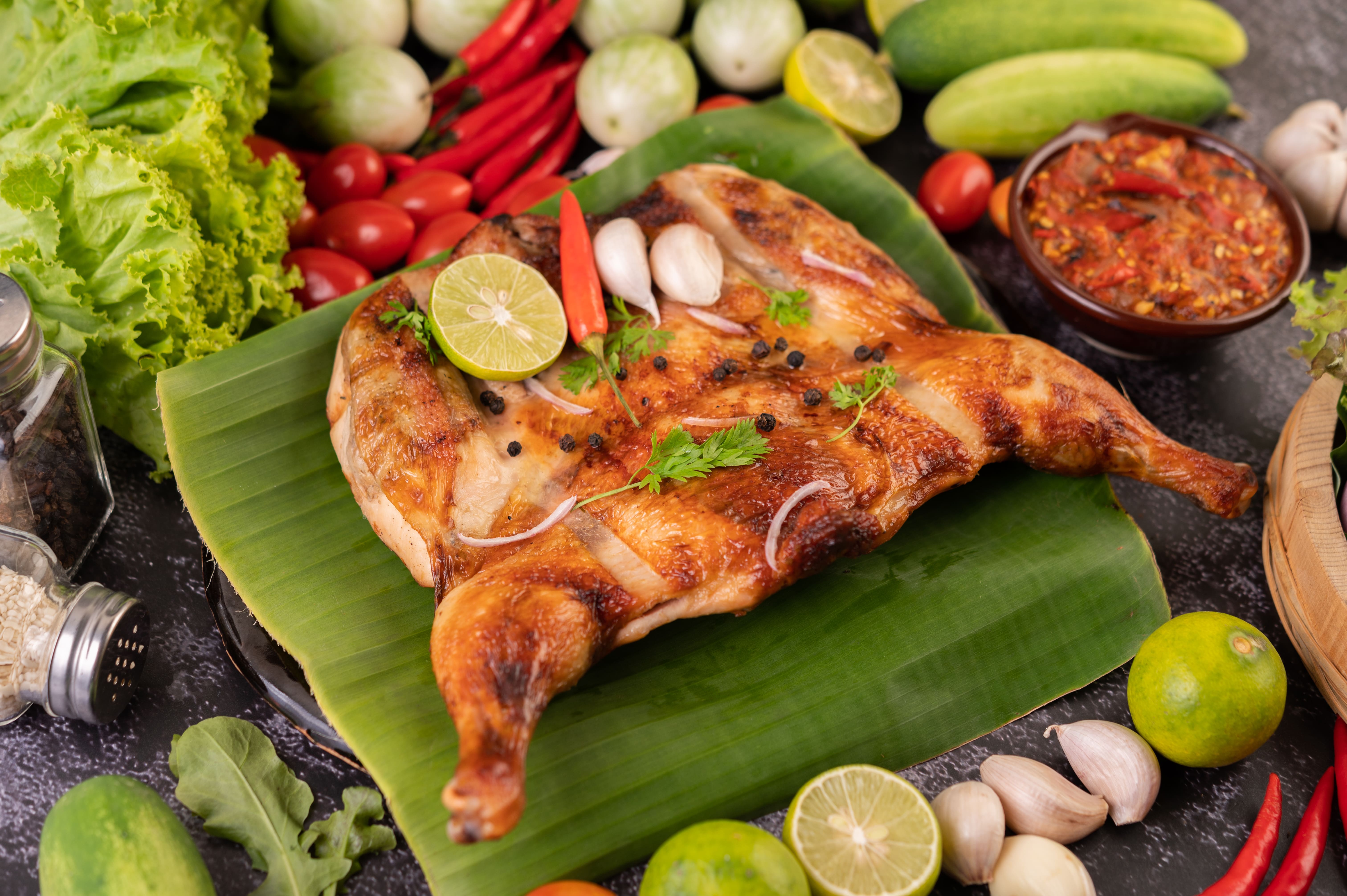 Ayam Taliwang, a spicy grilled chicken dish from Lombok, served on a banana leaf with fresh vegetables, limes, and sambal, available at warungs near Mawun Beach.