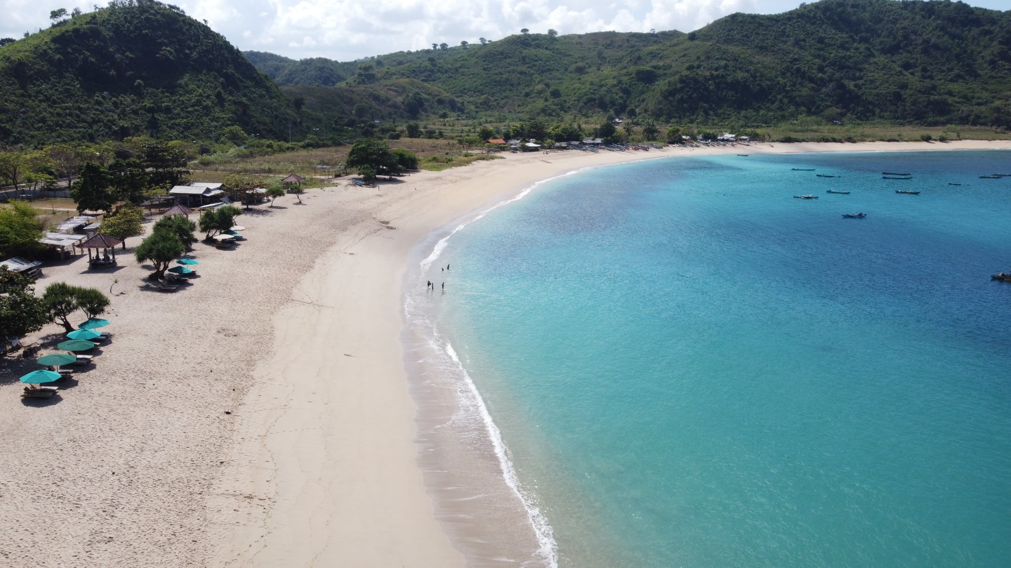 Aerial view of Mawun Beach in Lombok, Indonesia, featuring a crescent-shaped shoreline with golden sand, turquoise waters, and lush green hills in the background.