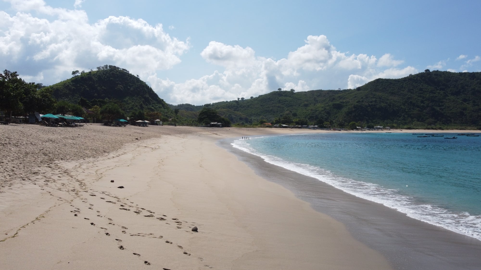 Ground-level view of Mawun Beach in Lombok, Indonesia, with golden sand, gentle waves, and green hills under a partly cloudy sky.
