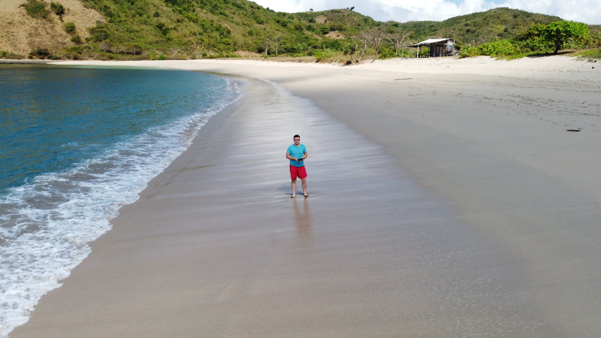 Person standing on the sandy shoreline of Mawun Beach in Lombok, Indonesia, with gentle waves and green hills in the background under a partly cloudy sky.