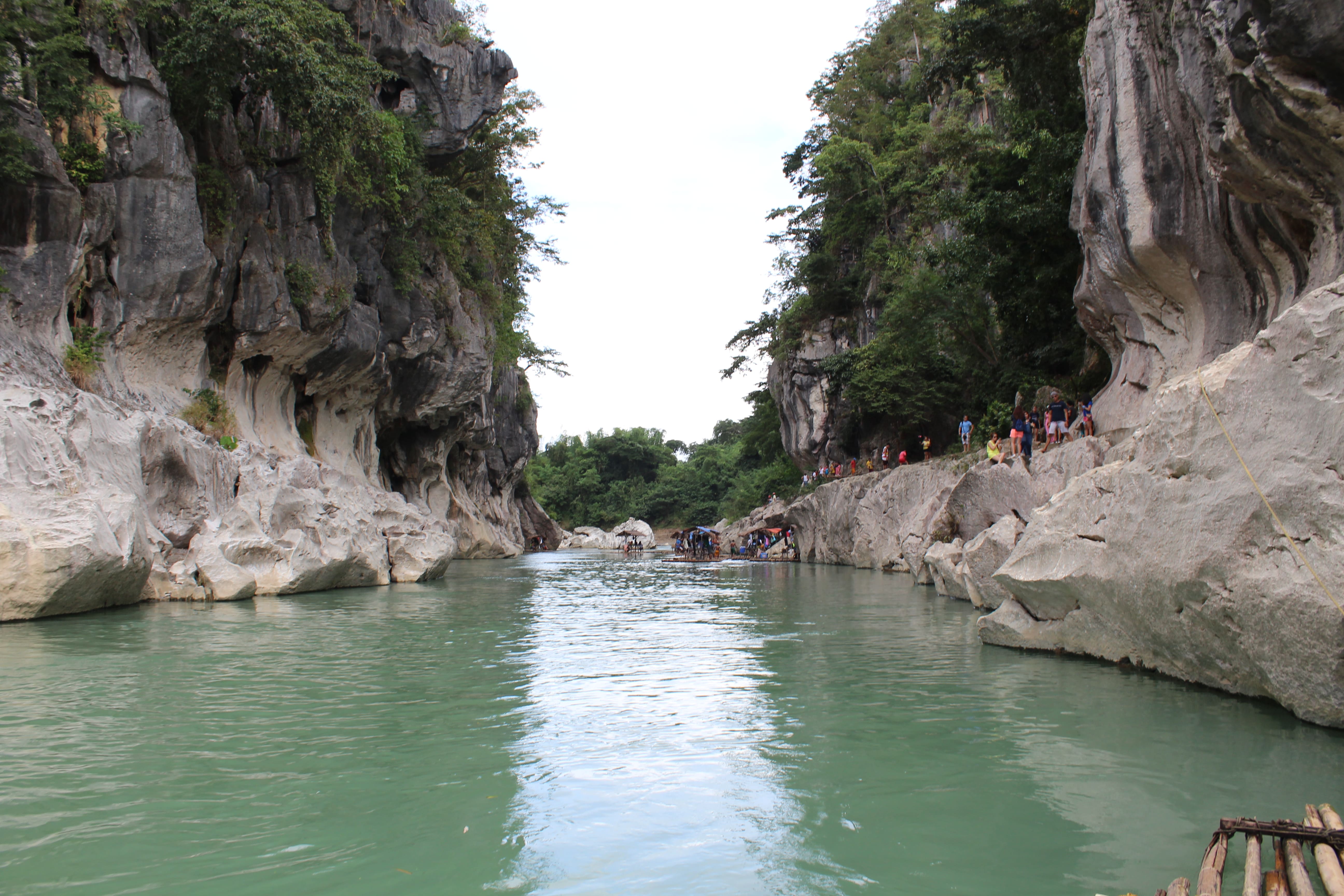 A scenic view of the Minalungao National Park and Peñaranda river in the Philippines. The image shows a calm, light green river flowing between tall, rocky limestone cliffs. Tourists are visible on the right side of the gorge, indicating recreational activity.
