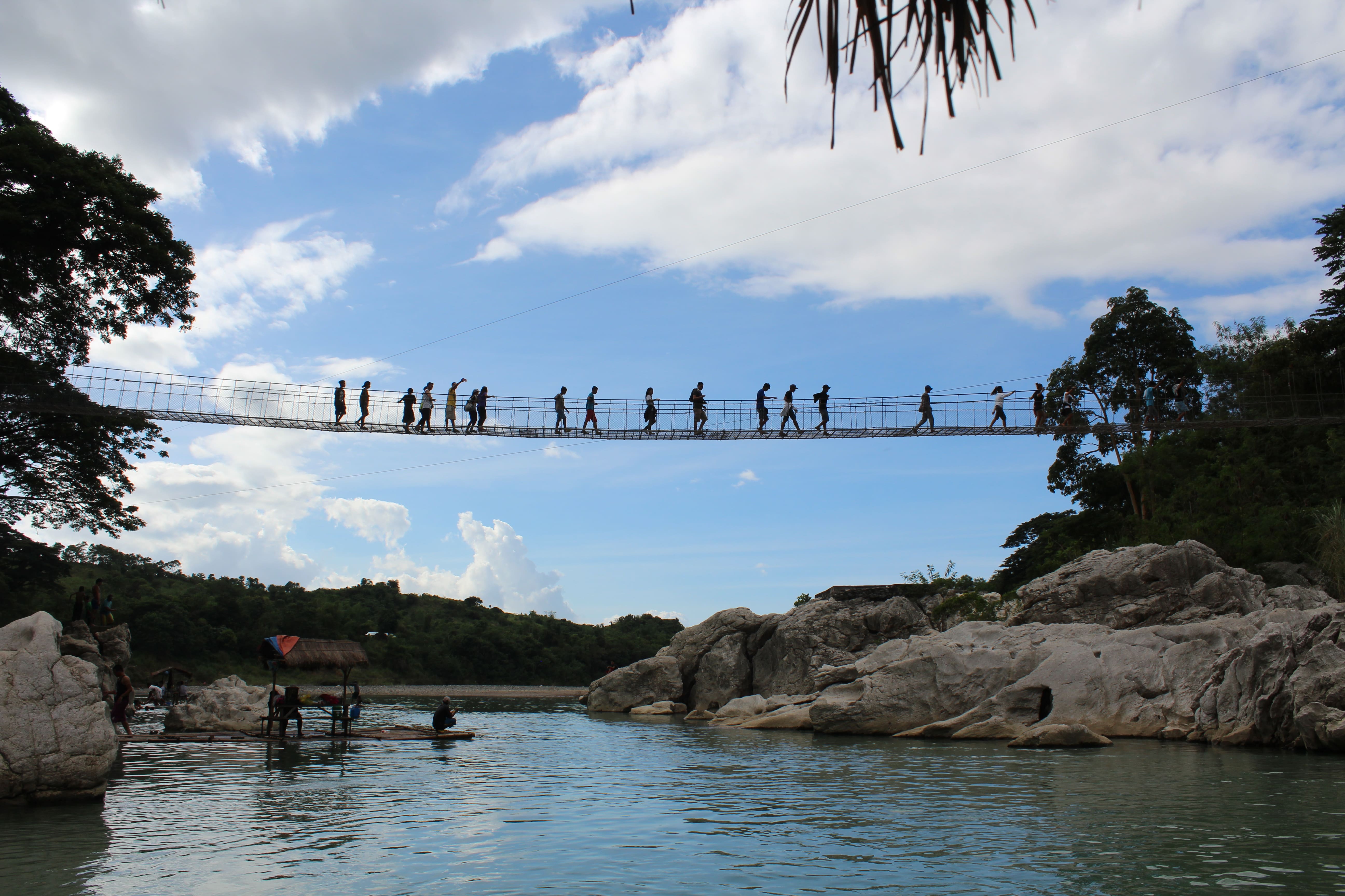 A group of people walking across a suspension bridge over the Peñaranda River at Minalungao National Park, with limestone cliffs and lush greenery under a partly cloudy sky, and a bamboo raft on the water below.