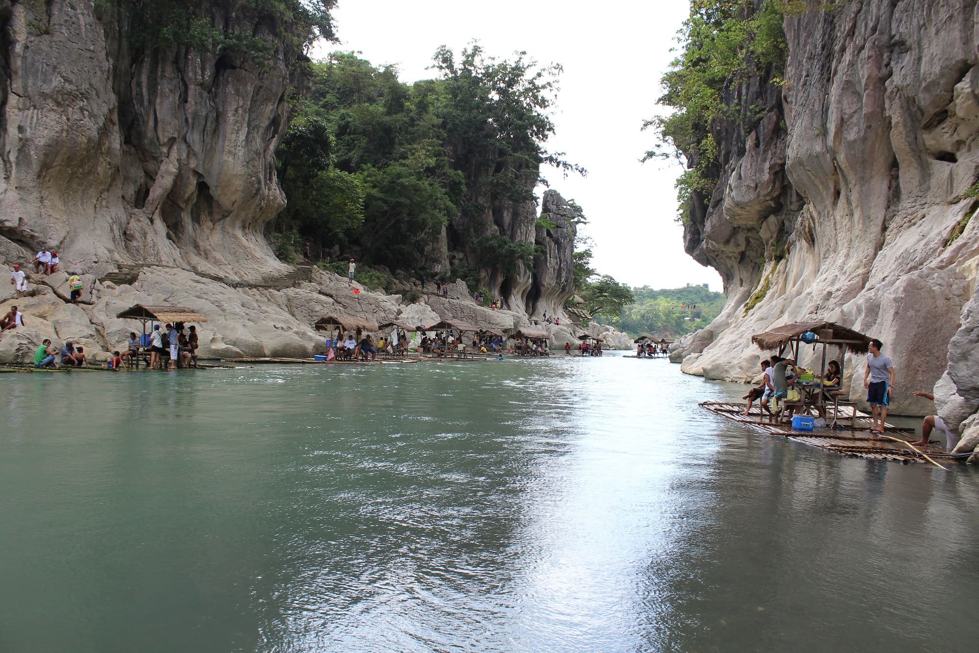 A wide shot of the Minalungao National Park, Philippines, featuring a river flowing between tall, light-colored limestone cliffs. People are gathered on bamboo rafts and small platforms along the riverbanks. Lush green trees grow atop the cliffs.