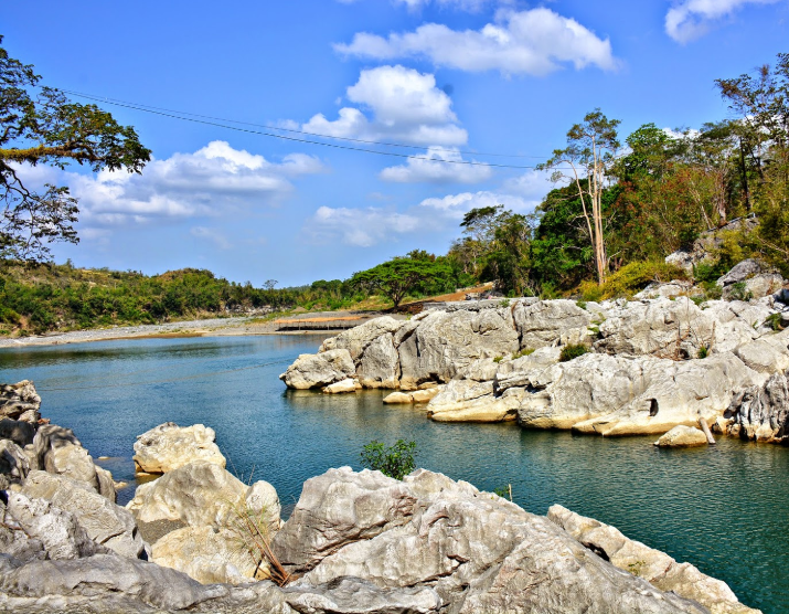 A scenic shot of the Peñaranda River in the Philippines, featuring a calm river with turquoise water surrounded by large, light-colored limestone rocks. Lush green trees line the riverbanks under a bright blue sky with scattered white clouds.