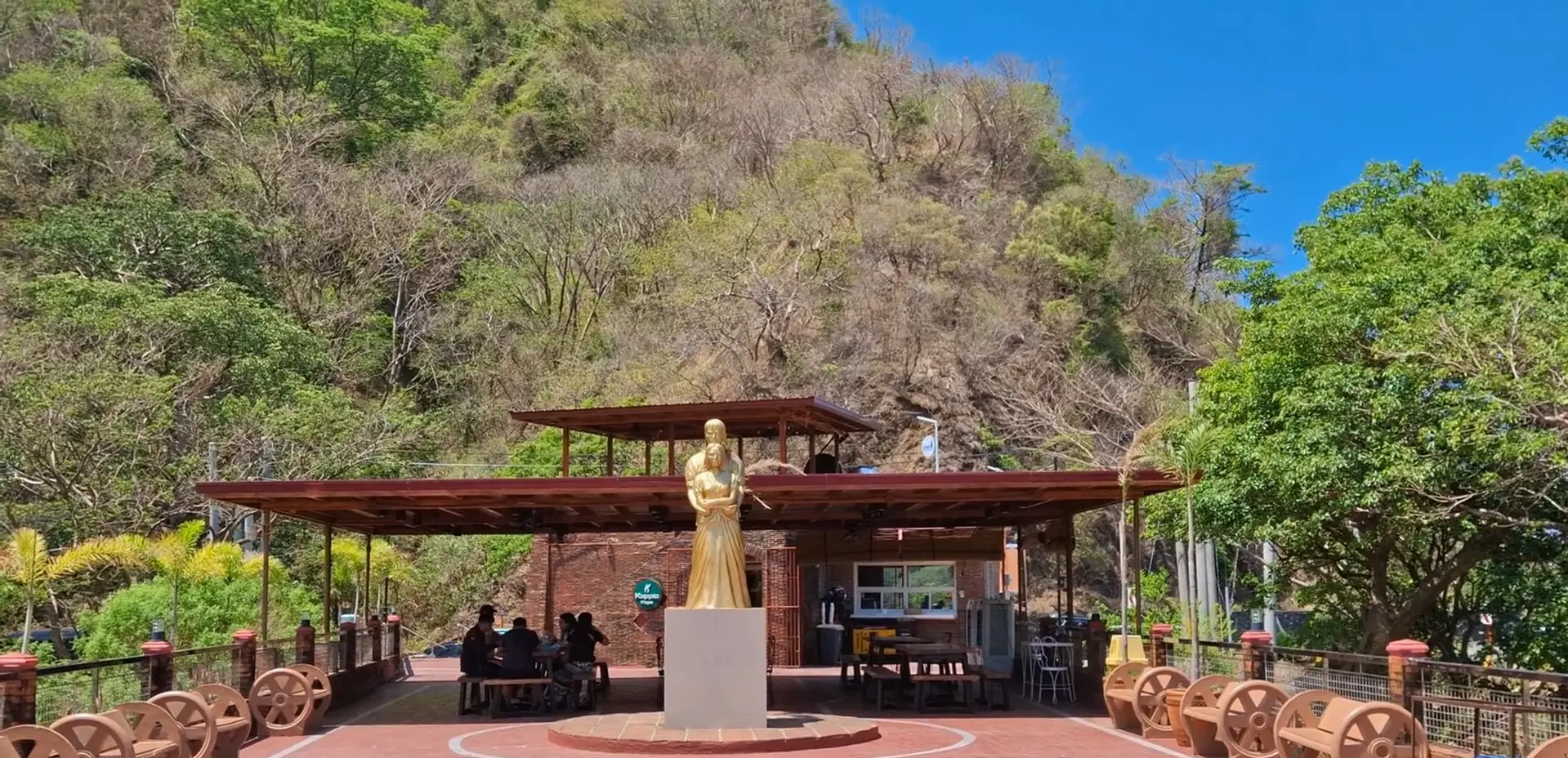 Golden statue of Gabriela Silang in a brick plaza against a steep, tree-covered hill.