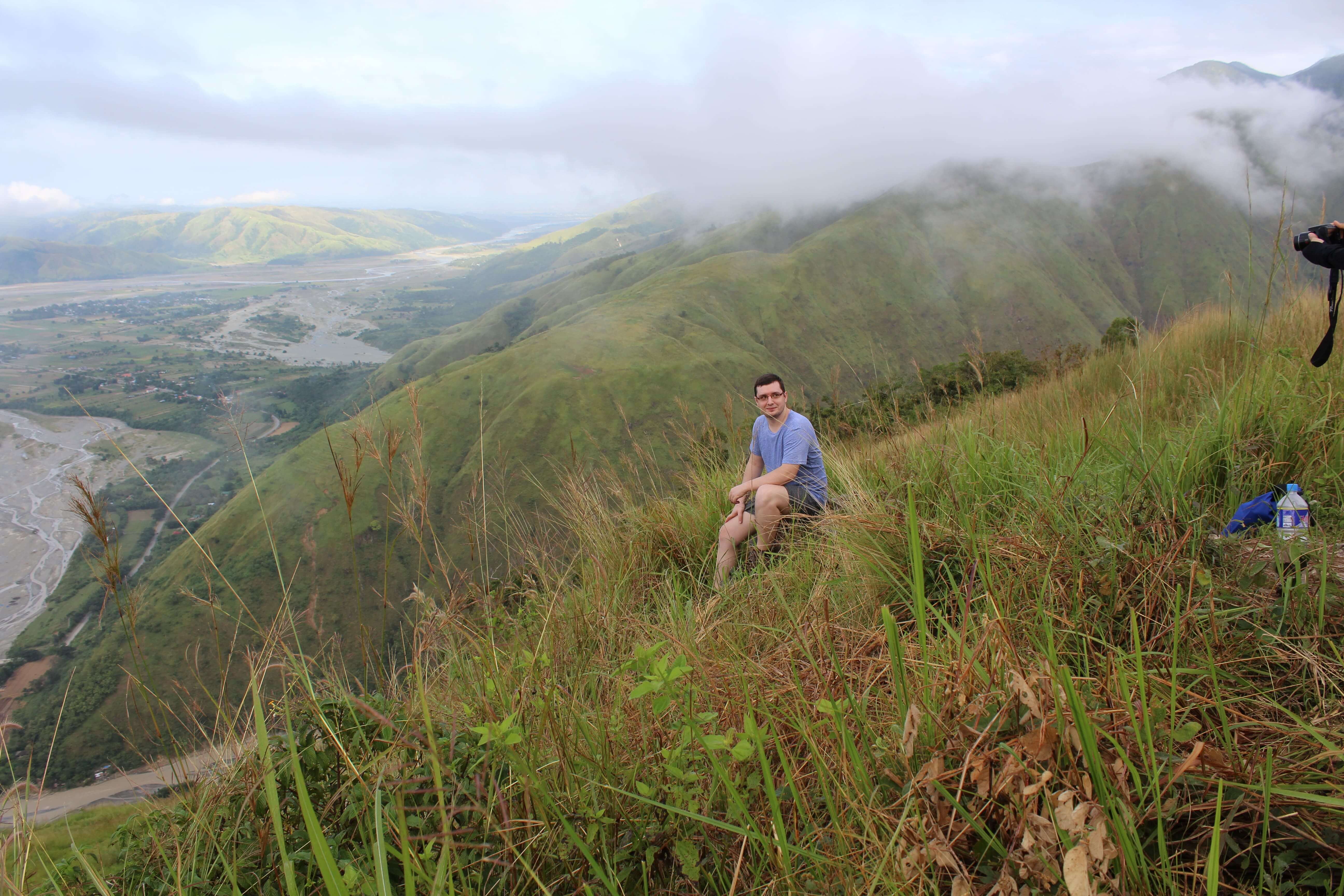 Hiker resting on Mount Sawi's grassy slope with misty mountains and valley view in the background