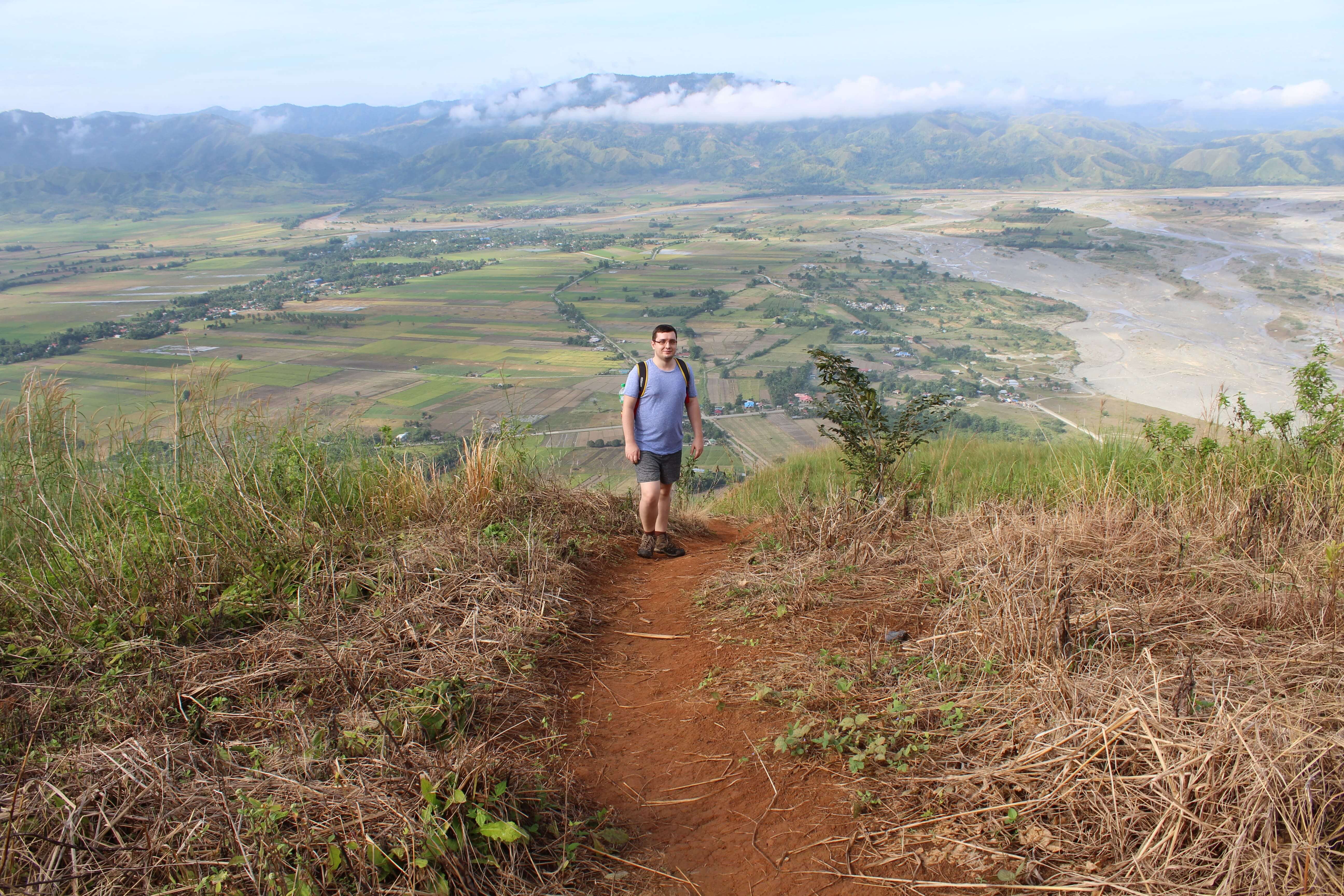 Hiker on Mount Sawi trail with tall grass, overlooking valley, river, and misty mountains