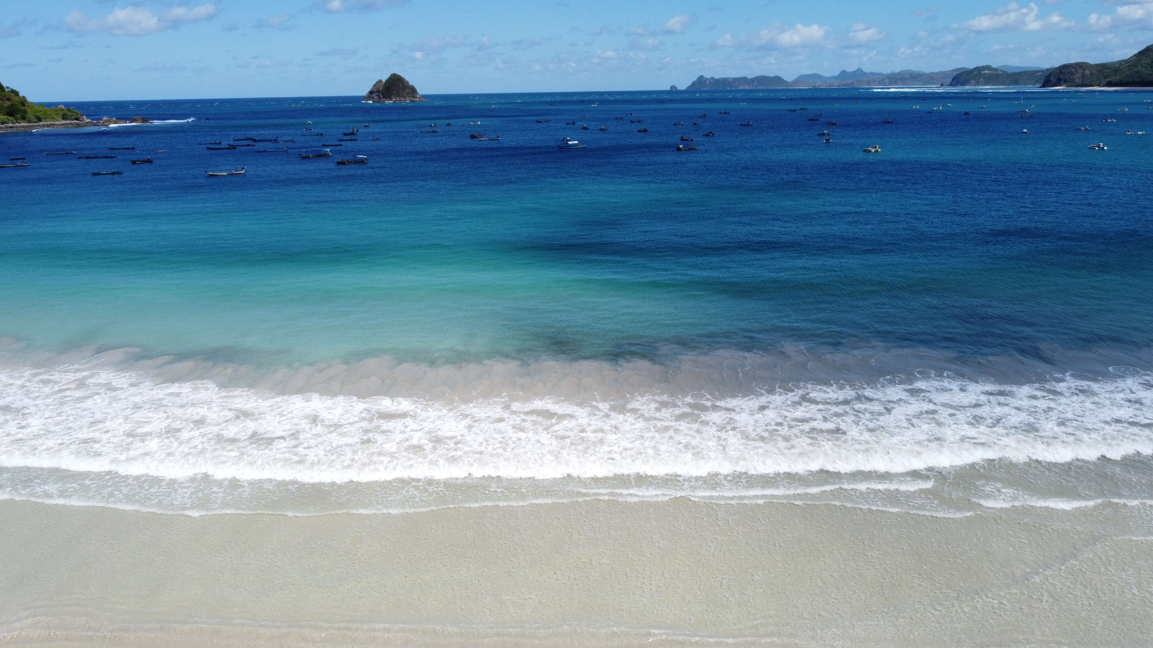 Aerial view of Selong Belanak Beach, Lombok, with white sands, turquoise and blue waters, boats, and distant green hills under a clear sky.