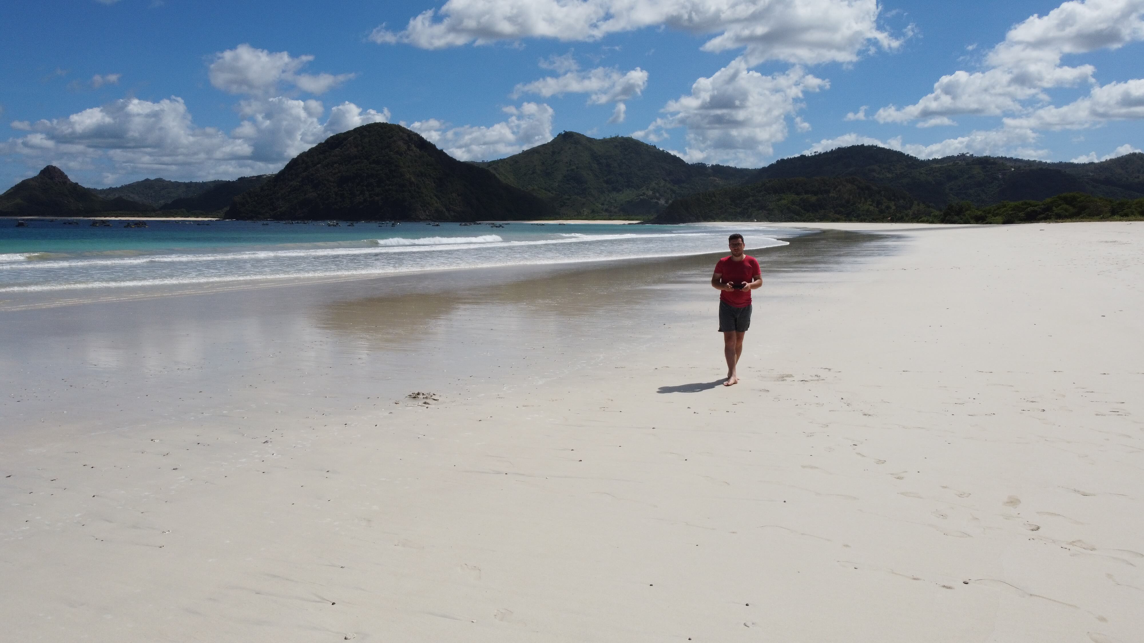 Scenic Selong Belanak Beach in Lombok, showcasing pristine shoreline, calm ocean with boats, and lush hills on the horizon.