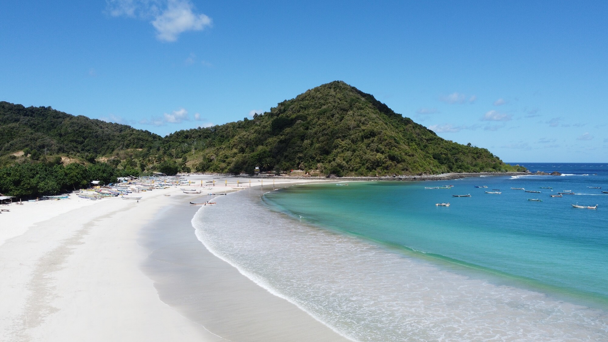 Aerial view of Selong Belanak Beach, Lombok, featuring white sand, turquoise waters, and lush green hills under a clear blue sky.