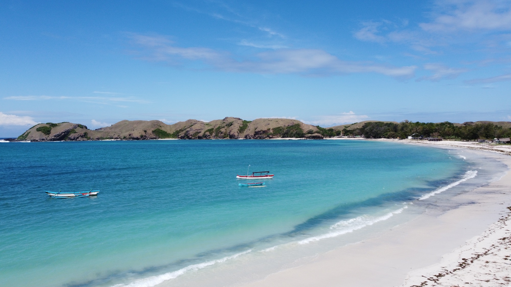 Stunning aerial view of Tanjung Aan Beach in Lombok, featuring pristine white sands, clear turquoise waters, and two traditional fishing boats floating peacefully. Lush green hills and a clear blue sky with scattered clouds frame the serene Indonesian beach paradise.