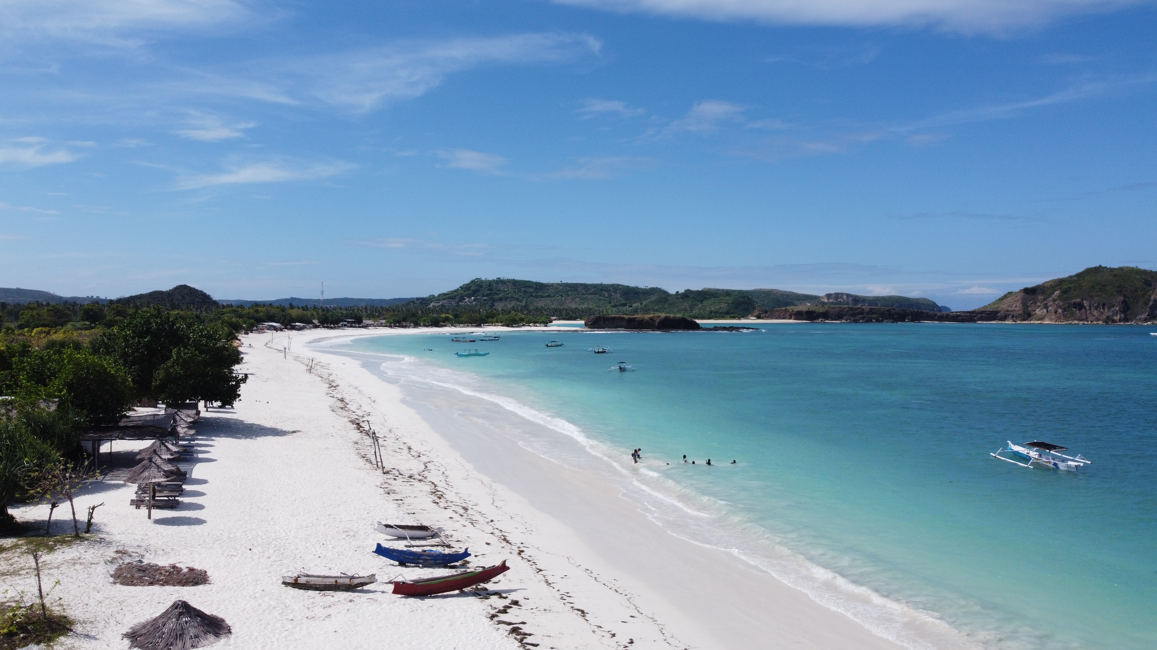 Breathtaking aerial view of Tanjung Aan Beach in Lombok, showcasing a long stretch of white sandy shore, crystal-clear turquoise waters, and traditional fishing boats. Thatched umbrellas line the beach, with lush green hills and a vibrant blue sky in the background, capturing the serene beauty of this Indonesian paradise.