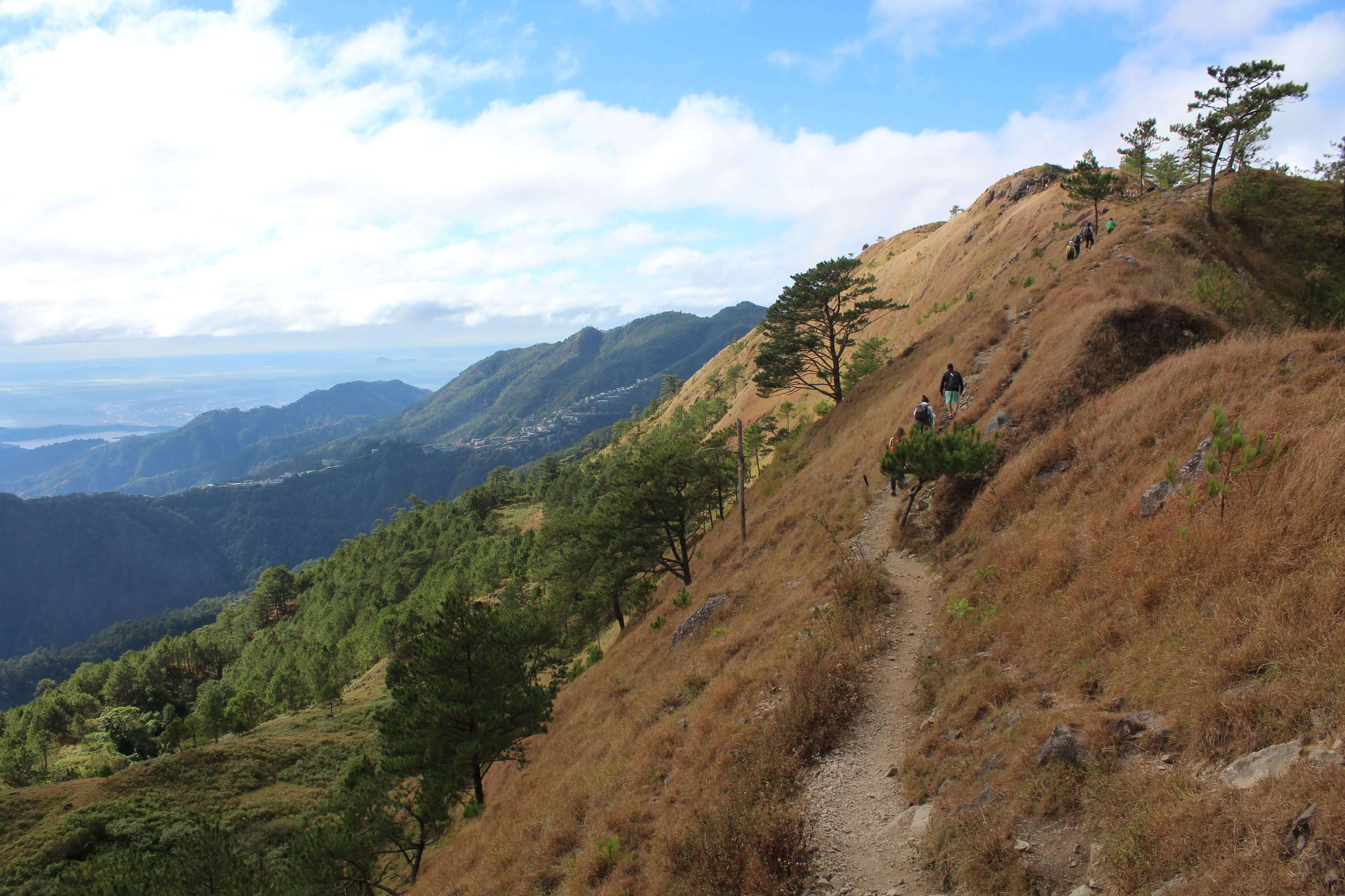 Hikers walking along the Mount Ulap trail in the Philippines, surrounded by pine trees, grassy slopes, and panoramic views of the Cordillera mountains.