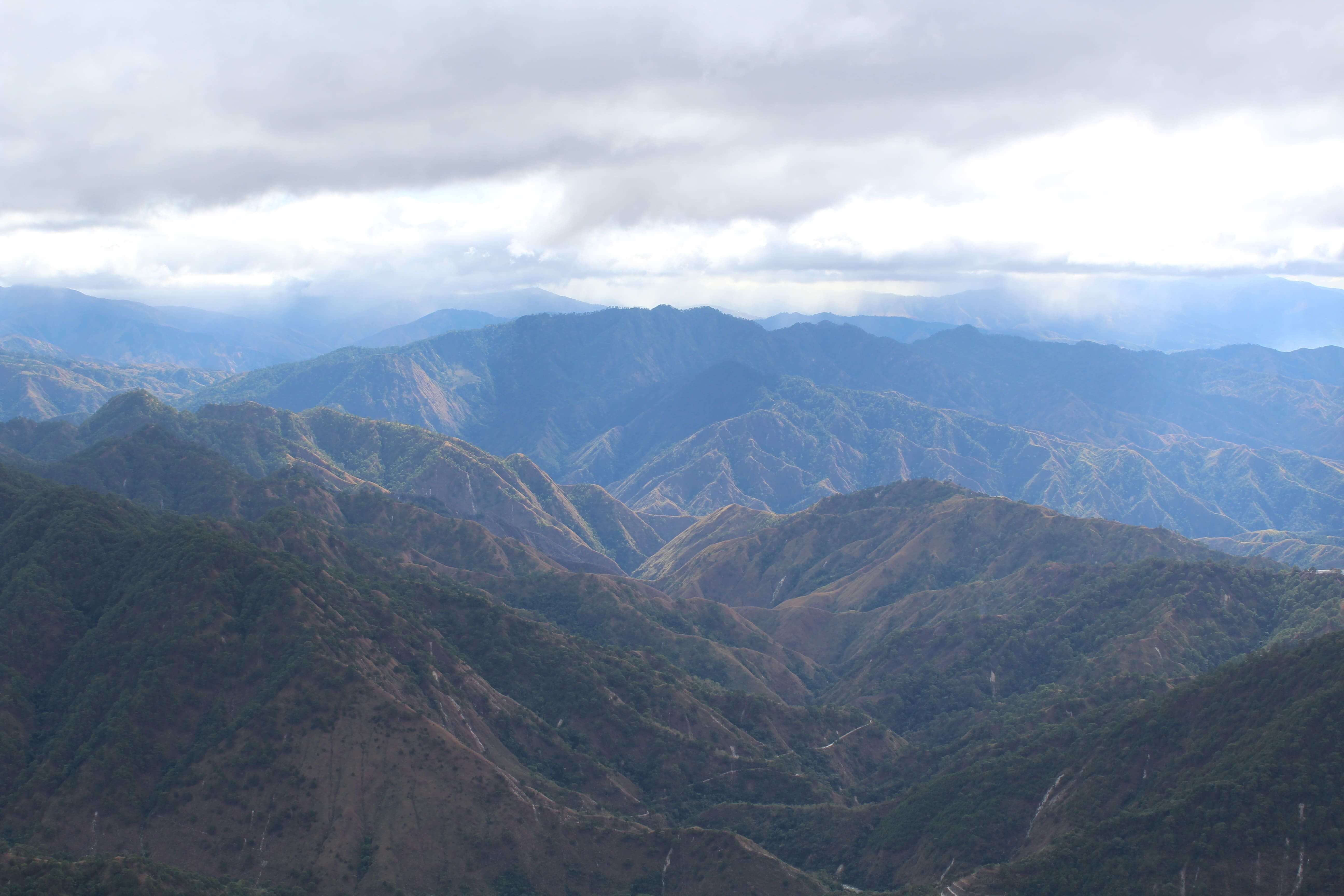 Aerial view of Mount Ulap’s landscape in the Philippines, featuring rolling hills, dense forests, and the Cordillera mountain range under a cloudy sky.