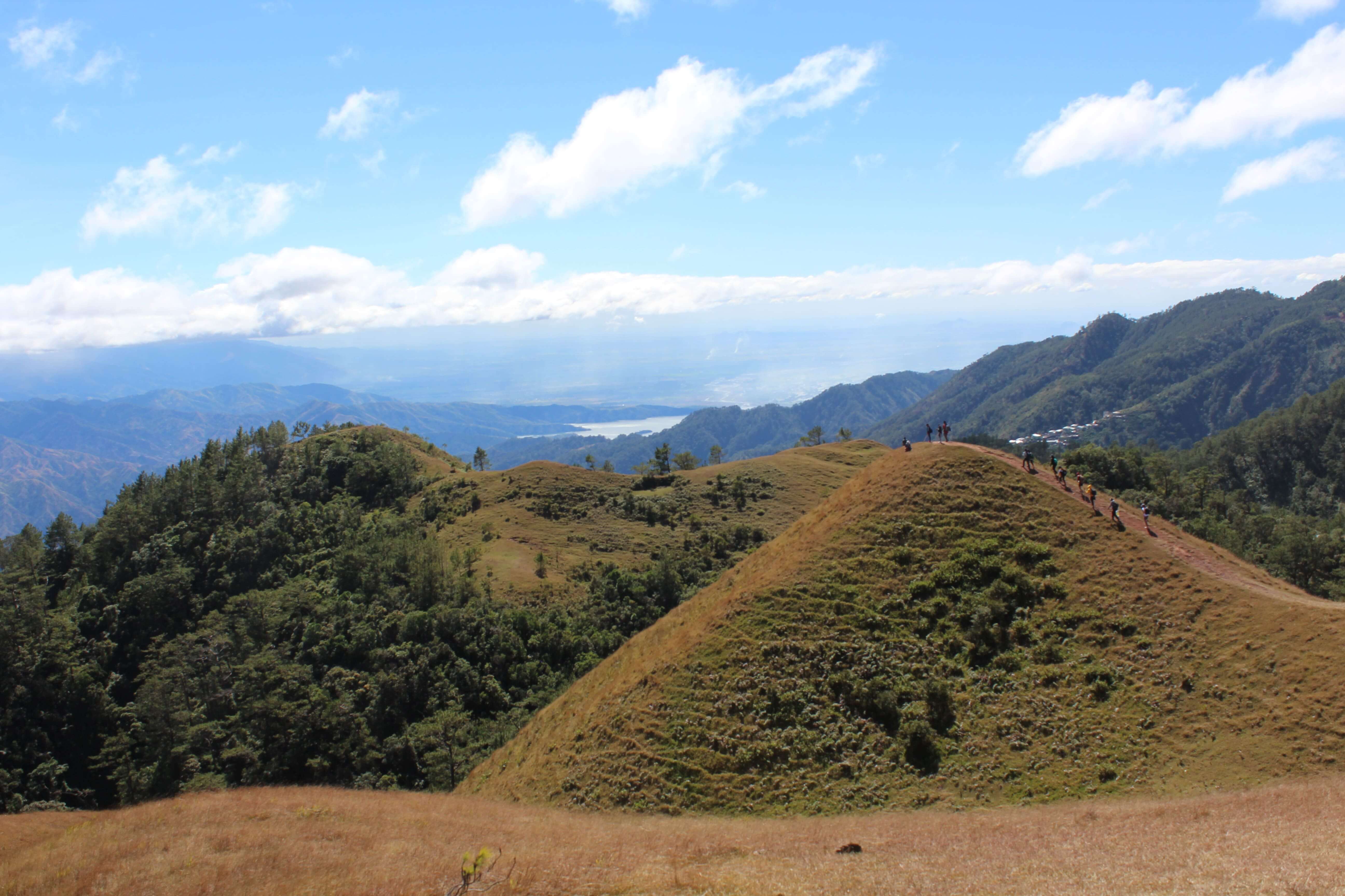 Hikers enjoying panoramic views along the Mount Ulap hiking trail in the Philippines, with rolling hills, lush forests, and distant mountains under a clear blue sky.