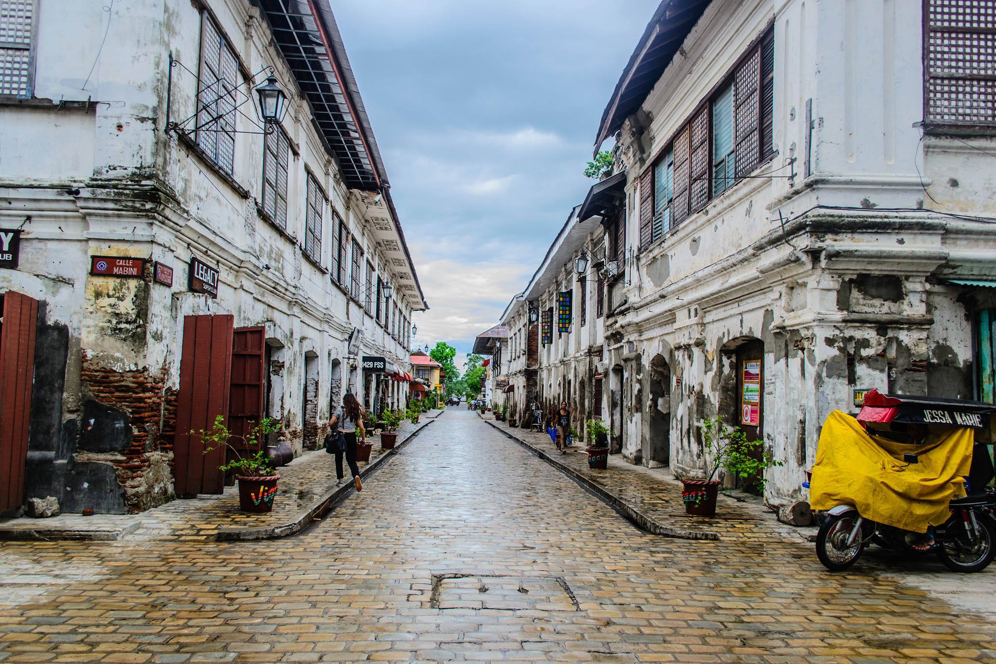 Historic cobblestone street in Vigan, Philippines, lined with old Spanish colonial buildings featuring capiz shell windows, potted plants, and a tricycle parked on the side, under a cloudy sky.