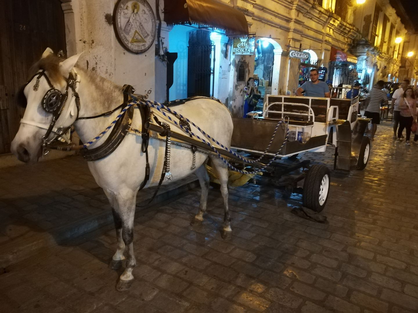 Horse-drawn carriage (kalesa) on a cobblestone street in Vigan, Philippines, at night, with a white horse harnessed to a cart, illuminated by streetlights, and historic buildings with shops in the background, people walking nearby.