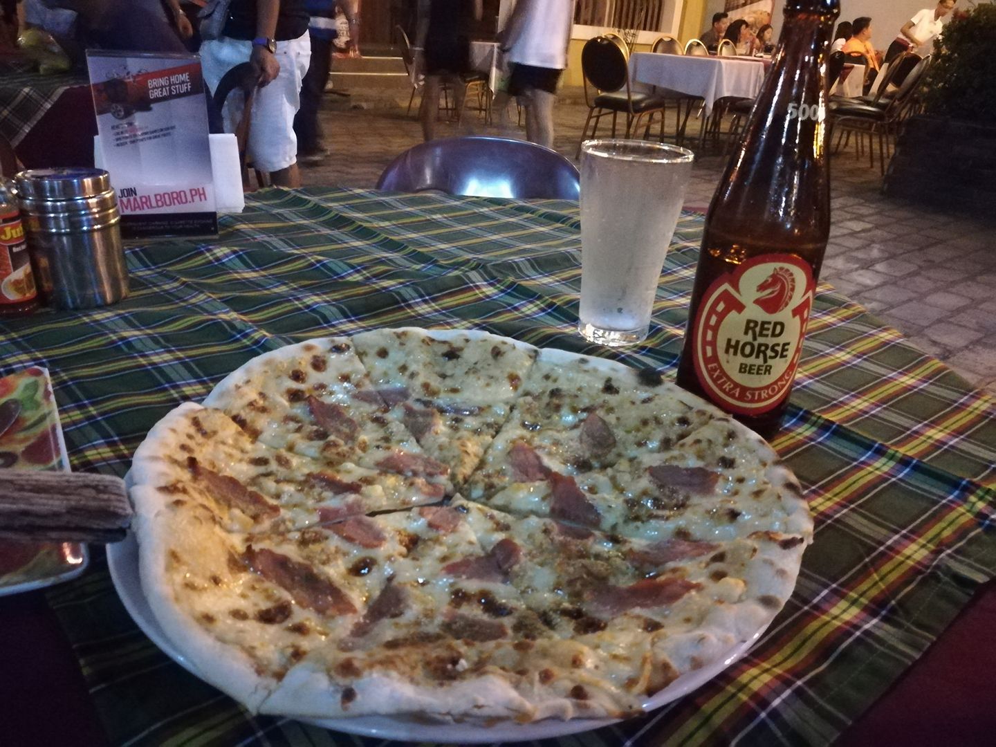Pizza with bacon and cheese on a white plate, served with a bottle of Red Horse beer and a glass of water on a plaid tablecloth, at an outdoor restaurant in Vigan, Philippines, with people dining in the background at night.